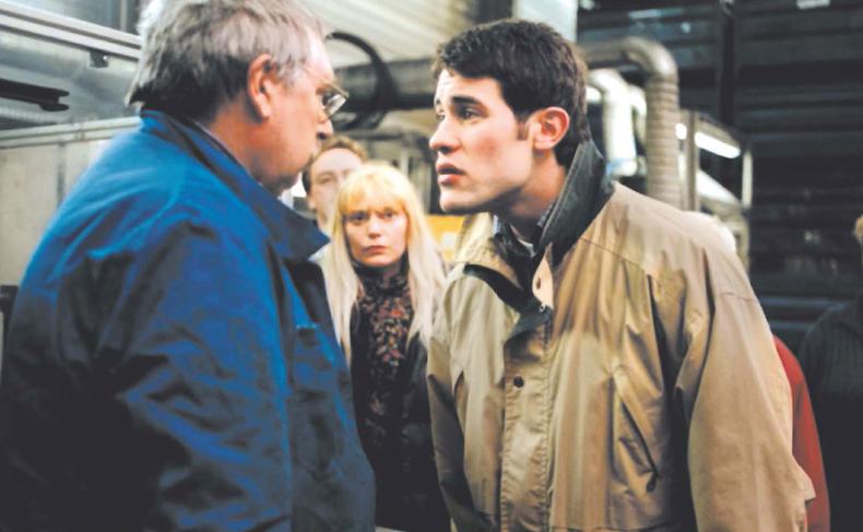 Still des Films RESSOURCES HUMAINES (F/UK 1999): In einer Fabrikhalle stehen zwei männlich gelesene Personen einander konfrontativ gegenüber. Im Hintergrund sehen mehrere Personen zu, darunter eine weiblich gelesene Person in der Bildmitte.